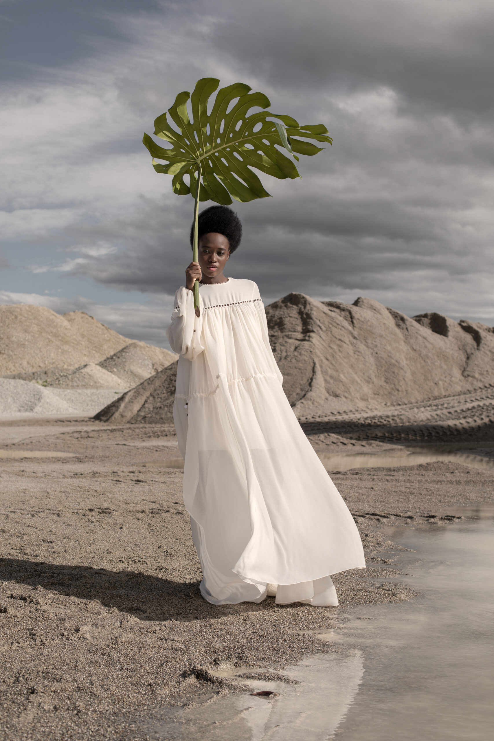 Fashion Stylist Sabine Holderer. Another Universe is a fashion editorial for La Botanica Magazine. The black model is standing in a sandy harbor, wearing a white long dress with few details by PHILOSOPHY DI LORENZO SERAFINI and shoes by LUCA DE FORTE. The model is holding a large green plate with a long handle in her right hand, as if she were holding an umbrella. Fashion Stylist Sabine Holderer Fleurs is a fashion editorial for l'official magazine. The photo is a closeup. The redhead model has her arm completely stamped with roses, like a tattoo. And she's wearing a pearl necklace.