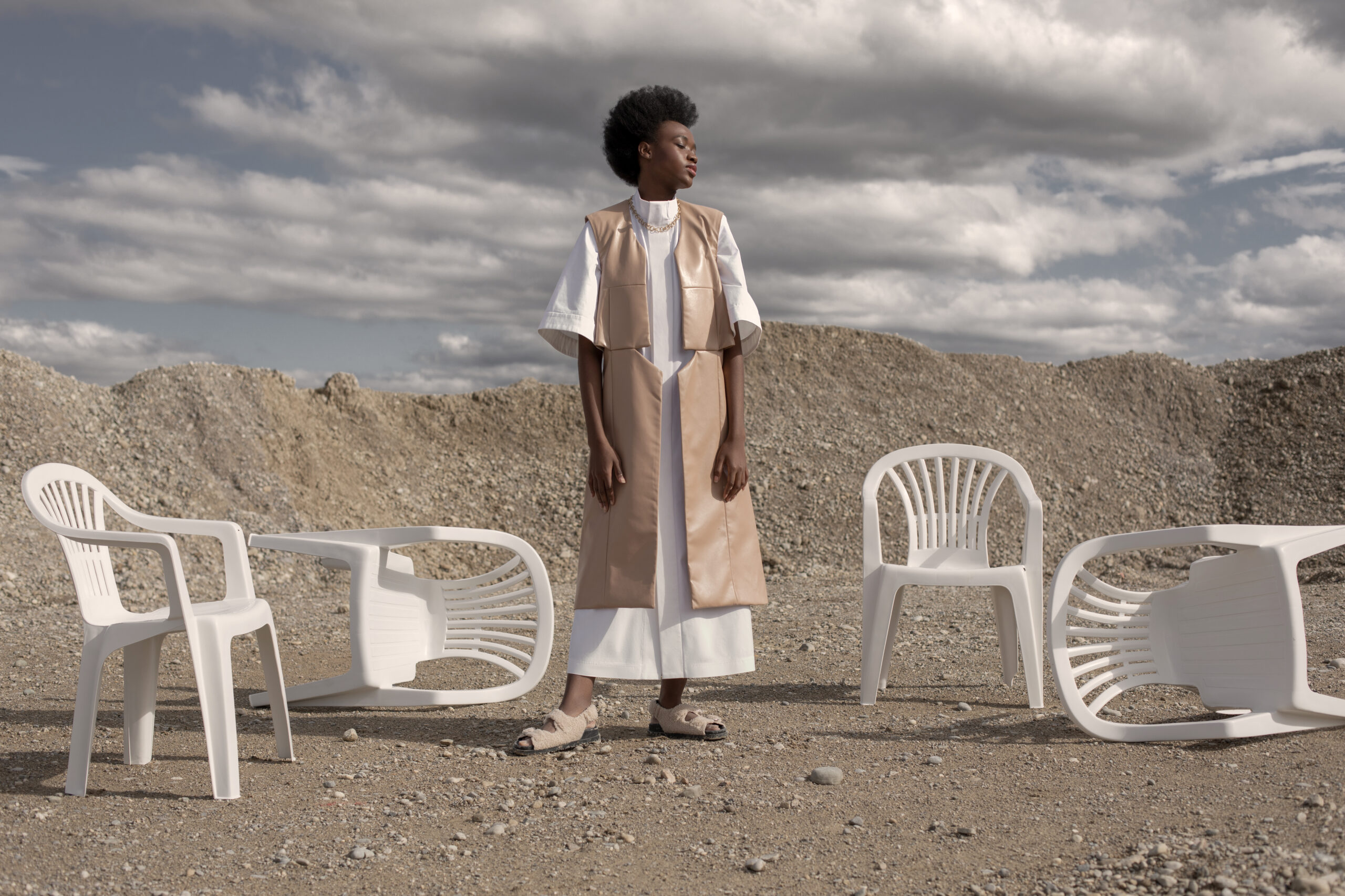 Another Univers is a fashion editorial for La Botanica Magazine. The black model is standing in a sandy harbor wearing a long plain white dress with wide sleeves and a long beige vest by LESC and shoes by Zara. Beside the model there are 4 white plastic chairs, 2 are standing and 2 are lying down. Fashion Stylist Sabine Holderer